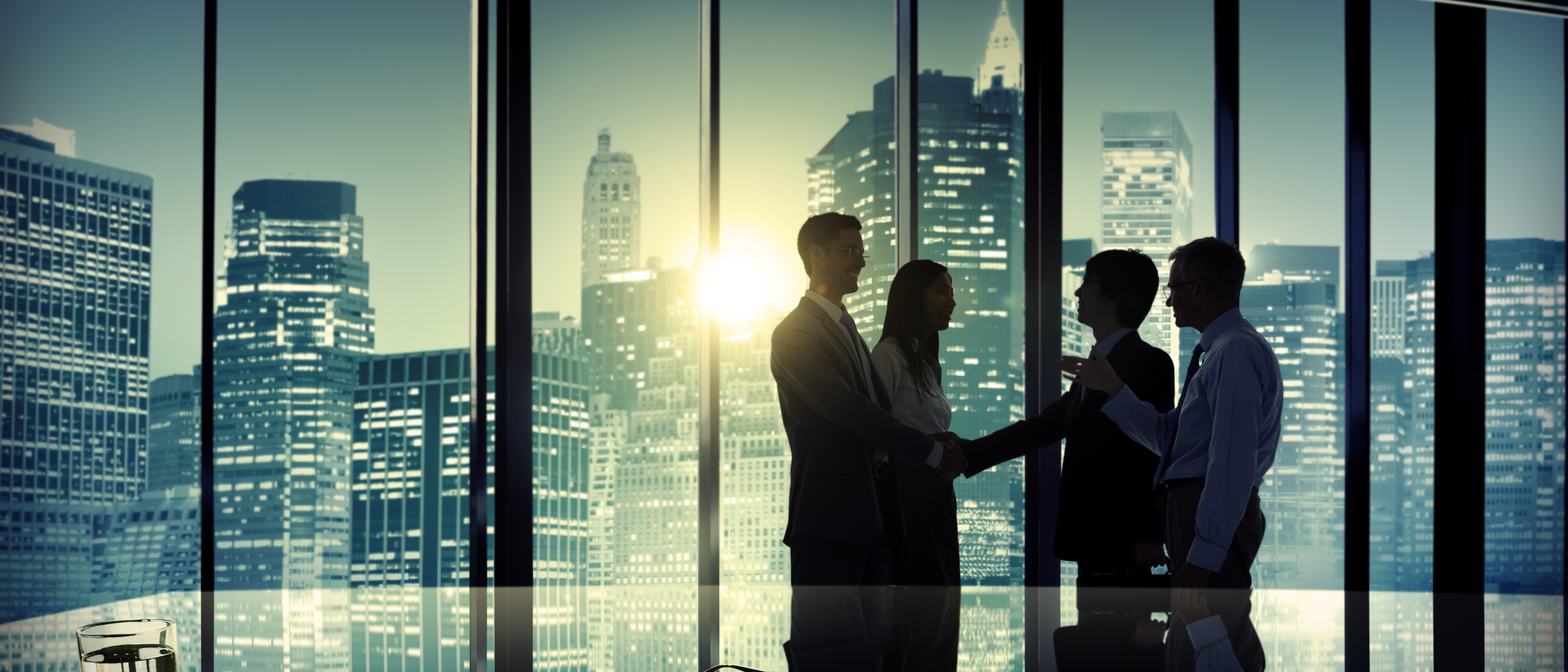 Silhouetted businesspeople shake hands in front of a large office window. 